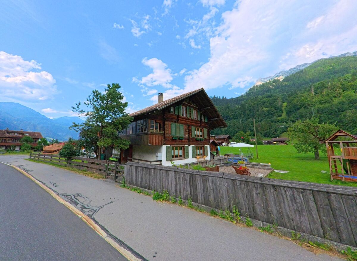 In Kandergrund (3716, Kanton Bern) steht ein traditionelles Holzhaus mit grünen Fensterläden am Rand einer Wiese, eingerahmt von bewaldeten Hängen und Bergkulisse