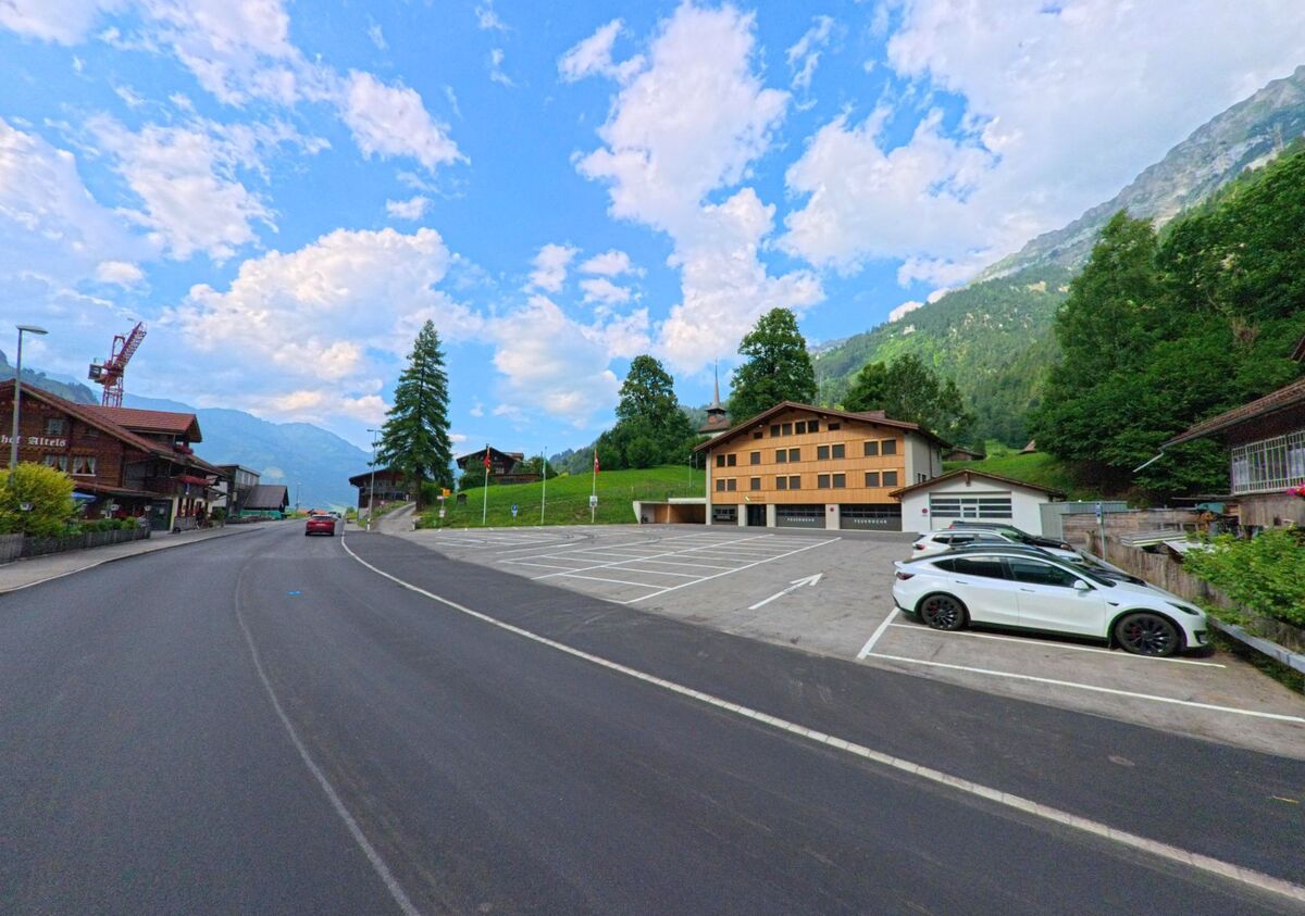 In Kandergrund (3716, Kanton Bern) liegt ein grosser Parkplatz vor einem modernen Holzbau, eingerahmt von steilen Hängen, Bäumen und angrenzender Dorfstruktur.