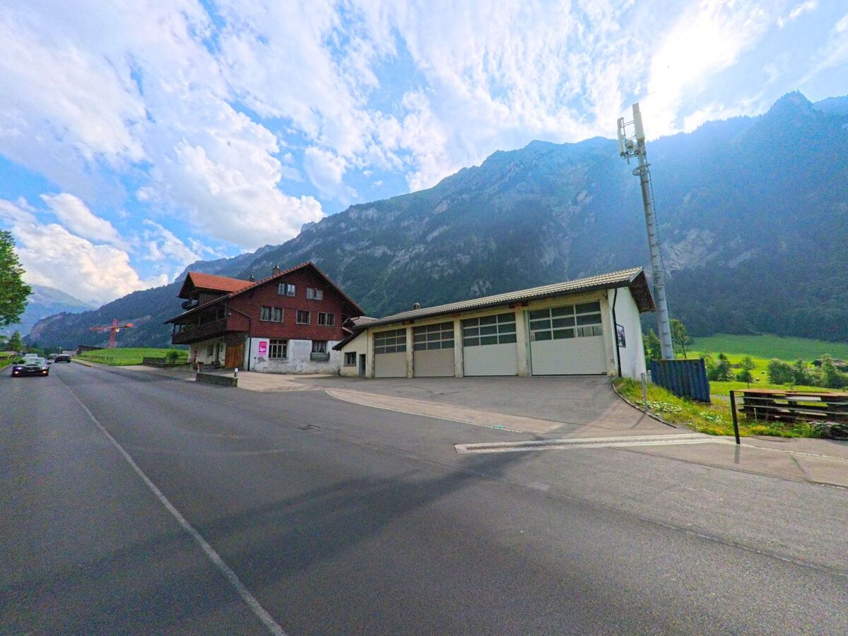 In Kandergrund (3716, Kanton Bern) stehen ein Wohnhaus und ein Gebäude mit Garagentoren an einer Hauptstrasse vor steil aufragenden Bergflanken und offener Landschaft.