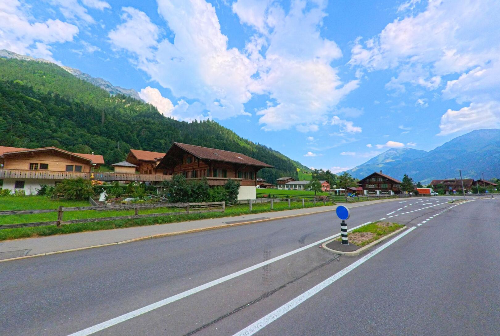 In Kandergrund (3716, Kanton Bern) verläuft eine breite Strasse mit Verkehrsinsel an einer lockeren Siedlung aus Chalets, umgeben von grünen Berghängen und weitem Himmel.
