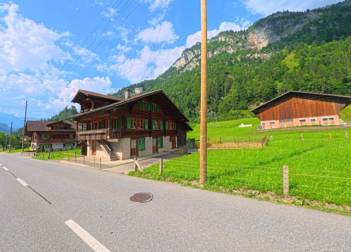 In Kandergrund (3717, Kanton Bern) säumen ein grosses Chalet und ein Wirtschaftsgebäude mit Holzfassade die Hauptstrasse, eingebettet in eine grüne Tallandschaft vor steiler Bergflanke.