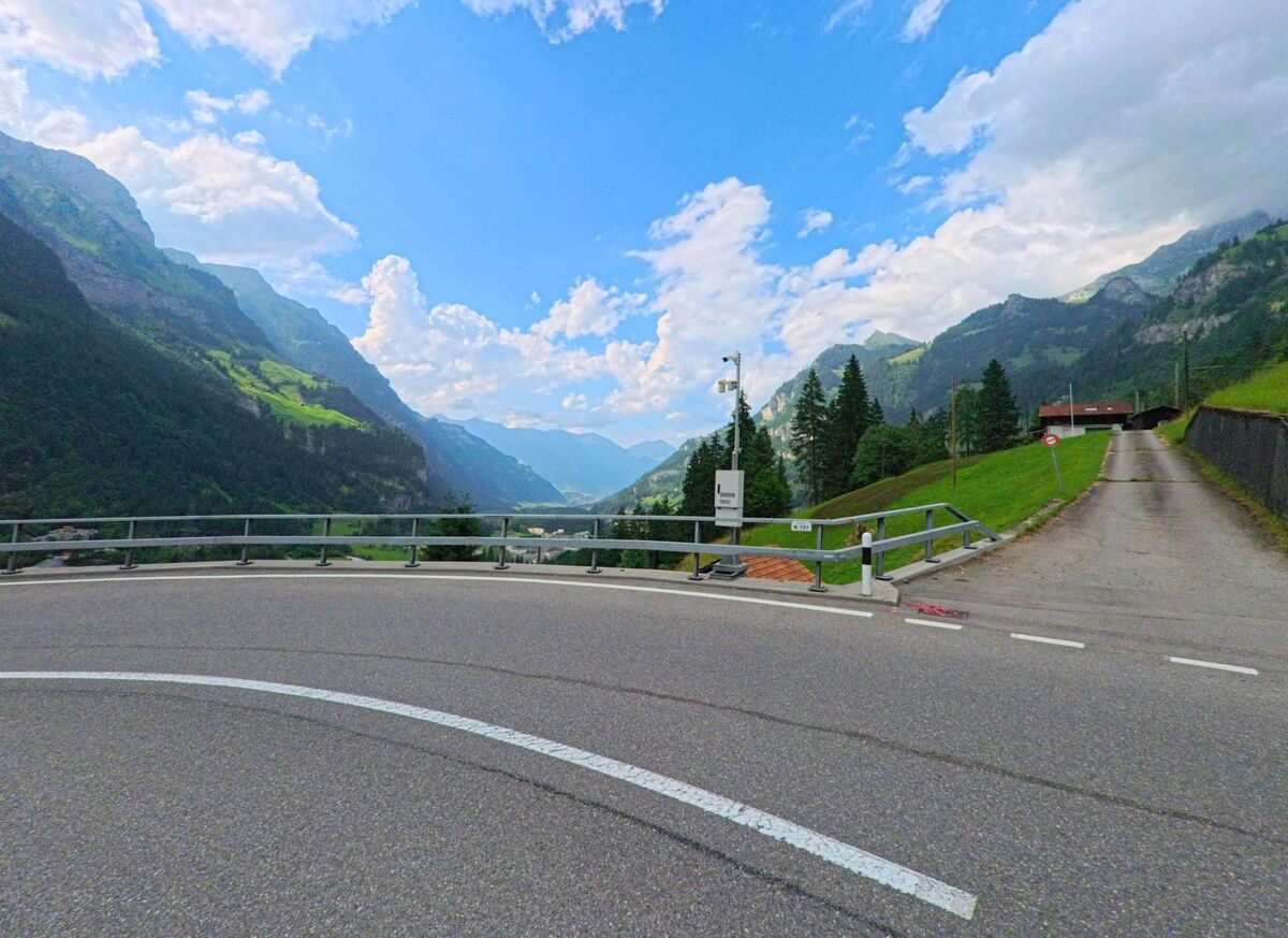 Von einer Kurve in Kandergrund (3717, Kanton Bern) öffnet sich der Blick ins tiefe Kandertal, eingerahmt von bewaldeten Berghängen und einer ansteigenden Seitenstrasse.