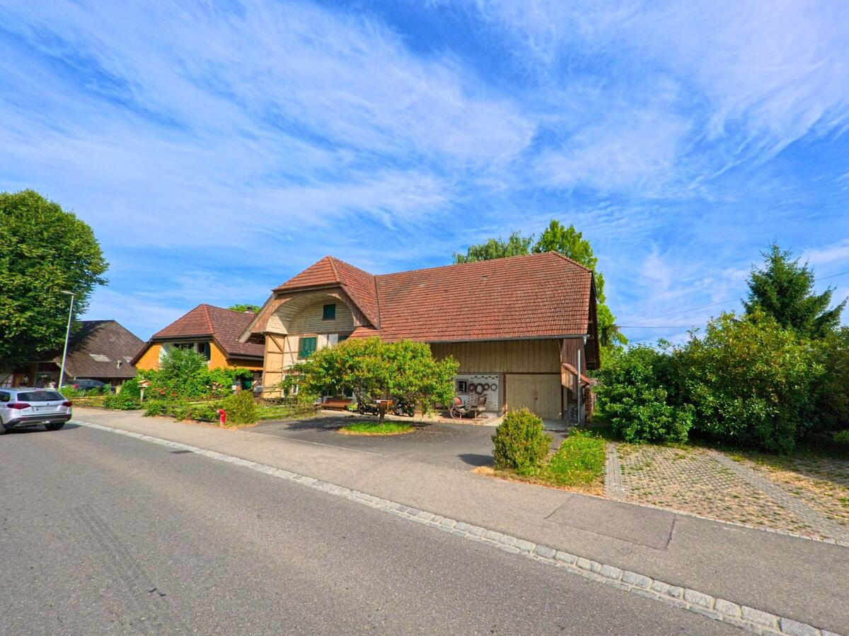 In einer ruhigen Seitenstrasse von Kappelen (3273, Kanton Bern) steht ein traditionelles Bauernhaus mit roten Ziegeldächern und gepflegtem Grün vor blauem Sommerhimmel.