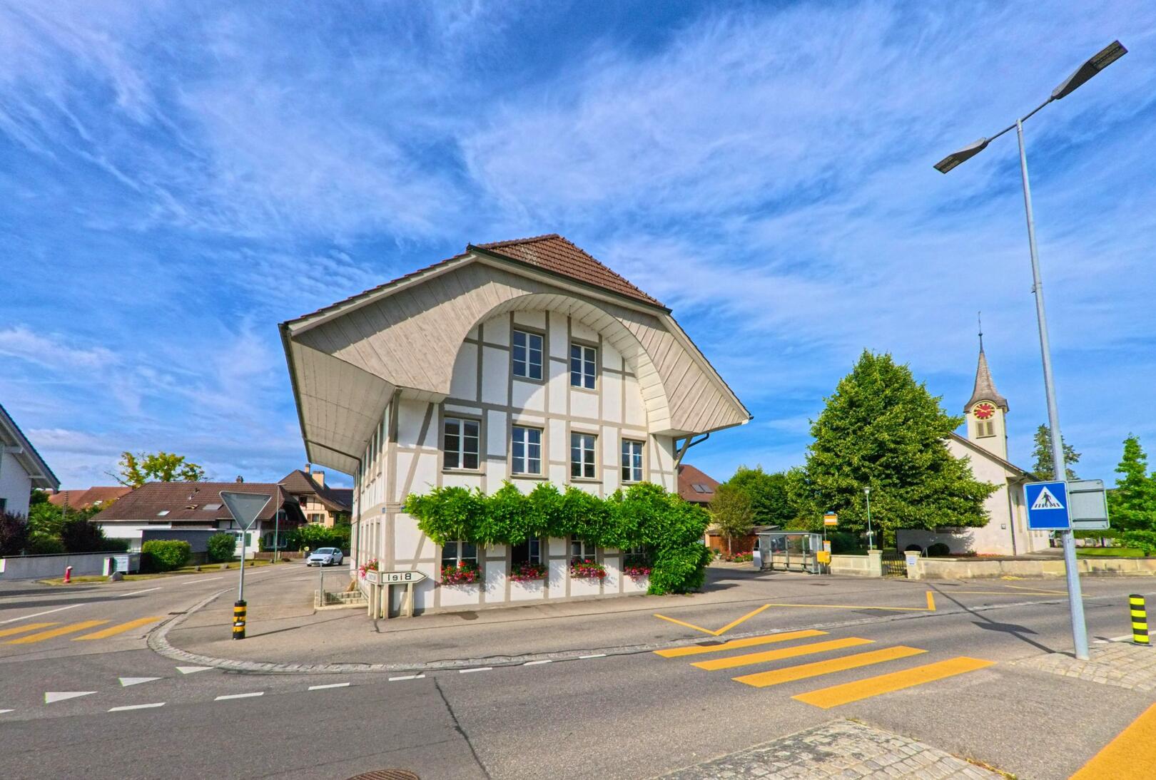 In der Gemeinde Kappelen (3273, Kanton Bern) steht an einer Kreuzung ein traditionelles Bauernhaus mit markanter Dachform, flankiert von der reformierten Kirche und einem Zebrastreifen im Vordergrund.