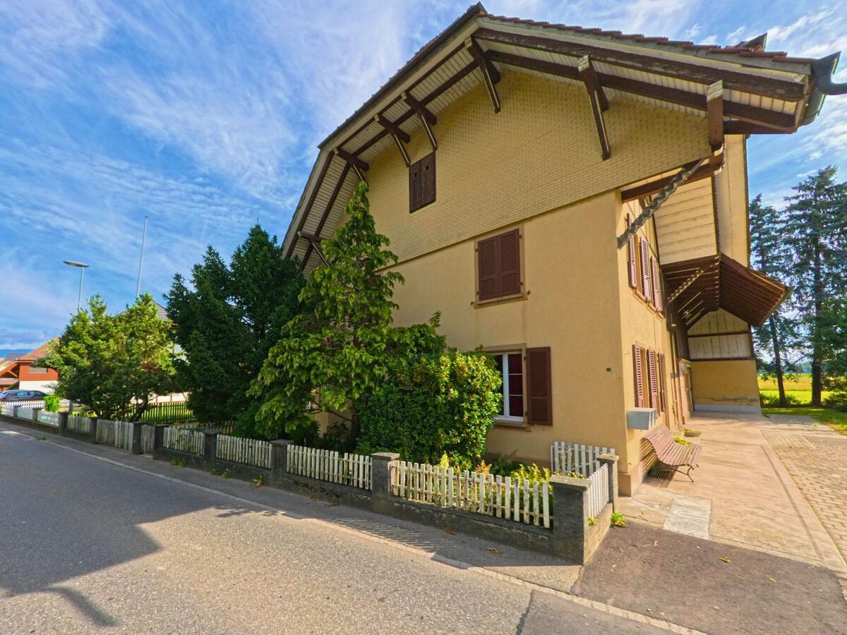 Ein gelb gestrichenes Haus mit markantem Dachvorsprung und Fensterläden prägt in Kappelen (3273, Kanton Bern) das Bild einer ruhigen Strasse am Rand grüner Felder.