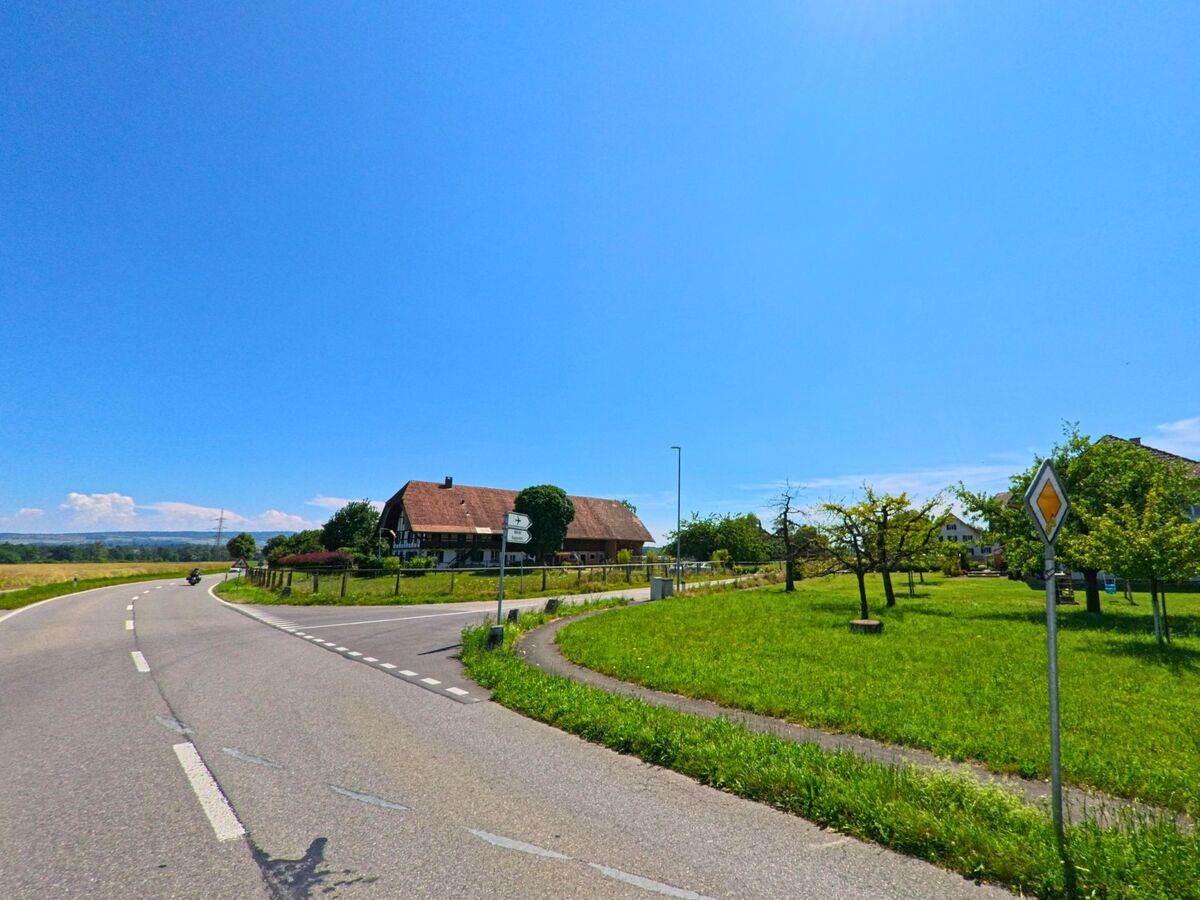 Am Ortseingang von Kappelen (3273, Kanton Bern) windet sich eine Landstrasse an einem grossen Bauernhaus vorbei durch grüne Wiesen und Obstbäume.