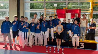 In Allschwil (4123, Kanton Baselland) präsentiert sich die Jugendgruppe von Karatekai Basel nach einem Wettkampf mit Medaillen und Pokalen in einer Sporthalle. Die jungen Karateka posieren gemeinsam mit ihren Trainern und Betreuern.
