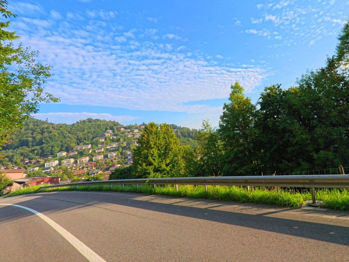 Von einer Strasse in Kehrsatz (3122, Kanton Bern) bietet sich ein Ausblick auf eine am Hang gelegene Wohnsiedlung, umgeben von Wald und grünen Huegeln.