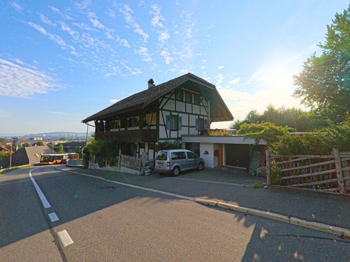Ein traditionelles Bauernhaus in Kehrsatz (3122, Kanton Bern) steht an einer abfallenden Strasse, waehrend die Morgensonne den Himmel erhellt.