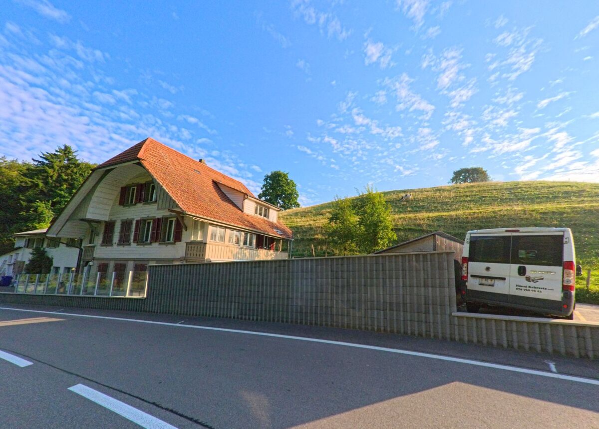 Vor einem sanft ansteigenden Huegel in Kehrsatz (3122, Kanton Bern) steht ein traditionelles Bauernhaus mit roten Fensterlaeden und grossem Walmdach.