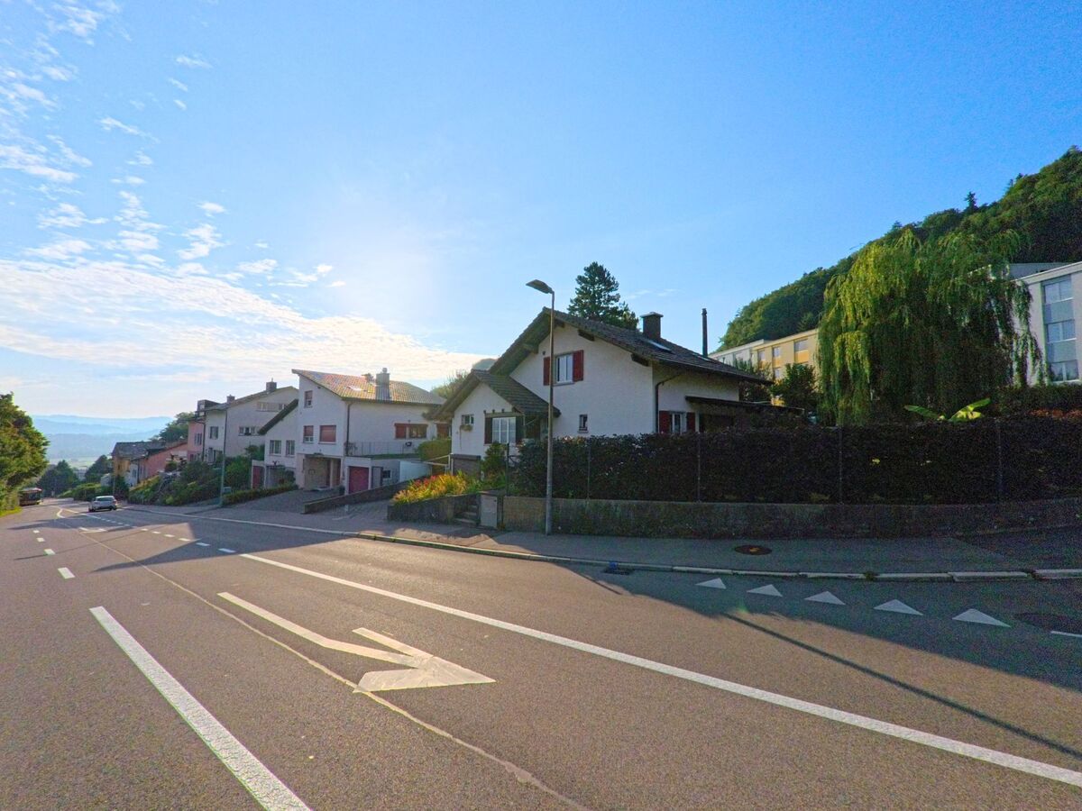 Entlang einer breiten Strasse in Kehrsatz (3122, Kanton Bern) stehen Einfamilienhaeuser mit Garagen, waehrend die Morgensonne tief am Himmel steht.