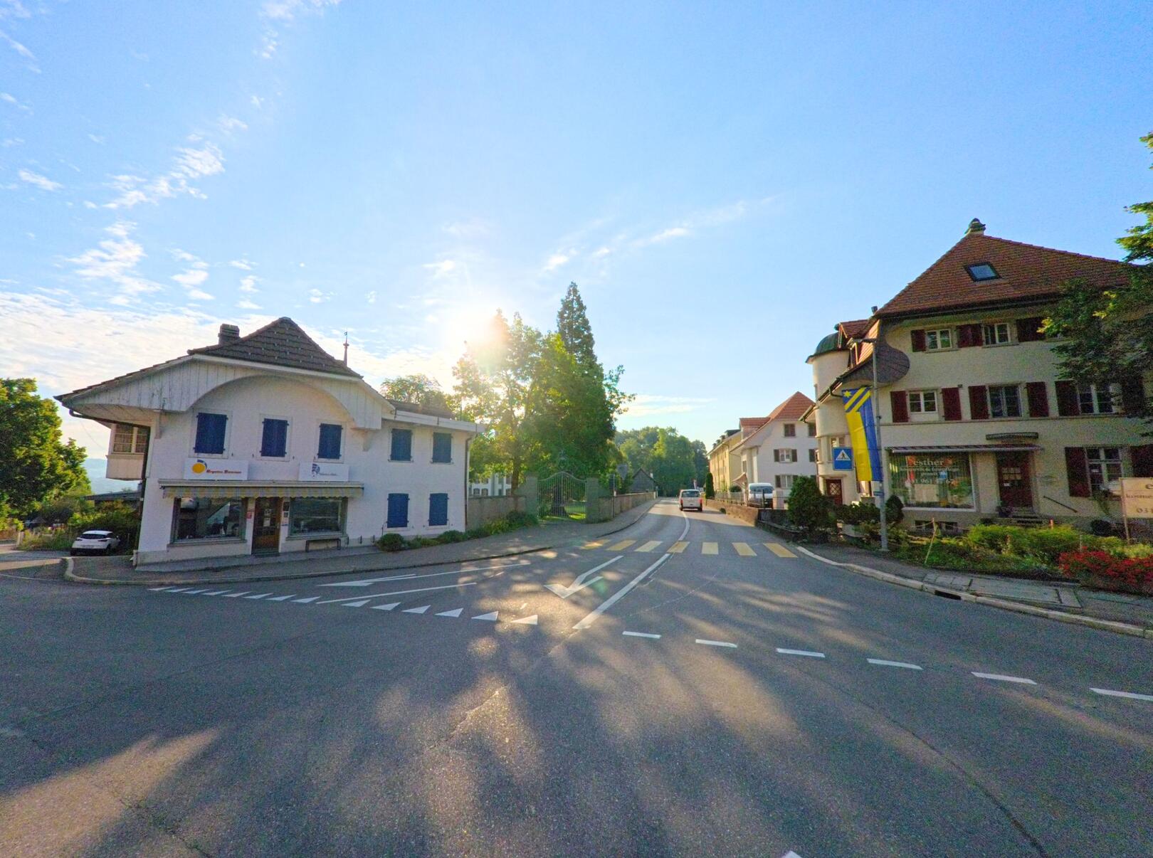 Am Morgen scheint die Sonne über die Hauptstrasse in Kehrsatz (3122, Kanton Bern), gesaeumt von Geschaefts- und Wohnhaeusern.