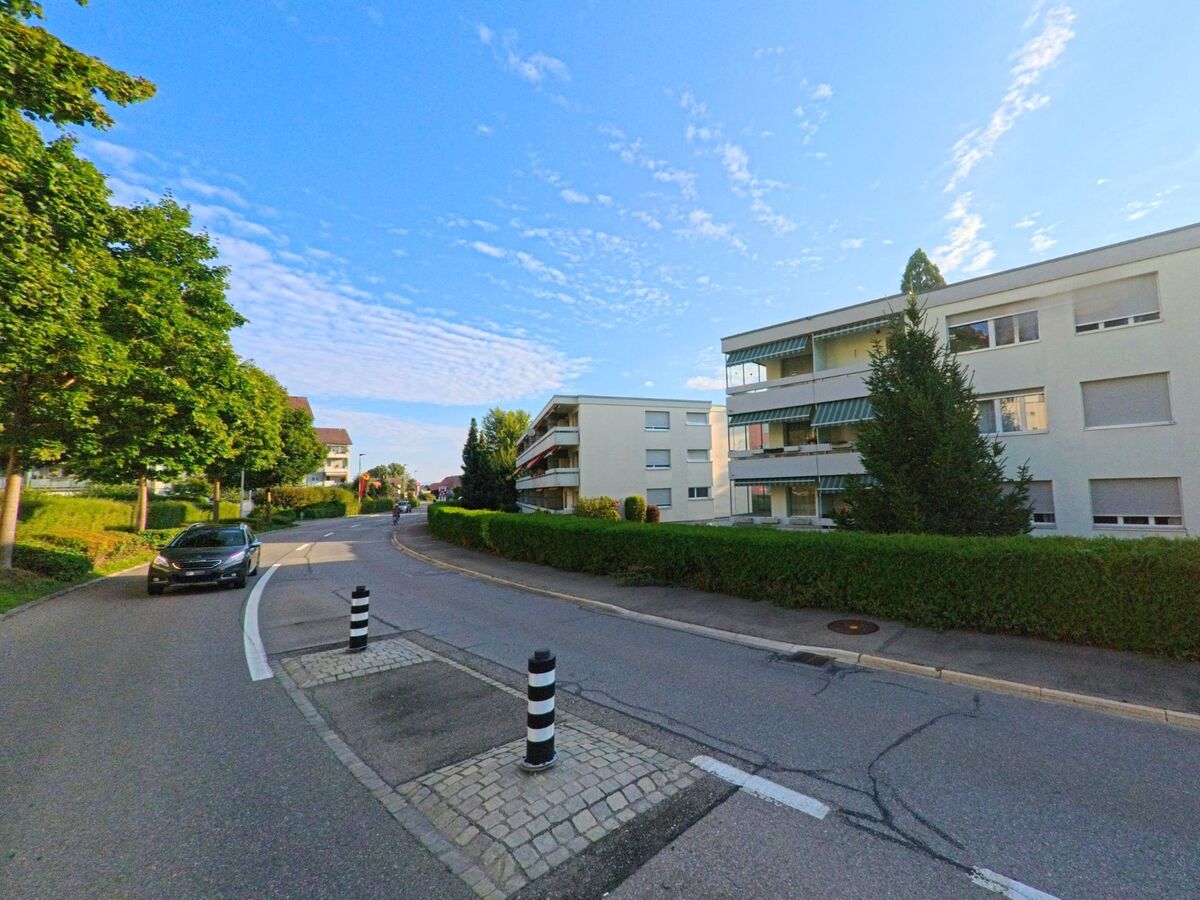 Mehrfamilienhaeuser mit grünen Markisen saeumen eine leicht geschwungene Strasse in Kehrsatz (3122, Kanton Bern), die von Baeumen gesaeumt wird.