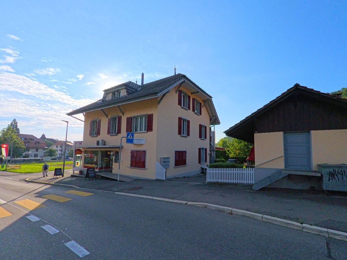An einer Strassenecke in Kehrsatz (3122, Kanton Bern) steht ein Gebaeude mit roten Fensterlaeden und einem kleinen Geschaeft im Erdgeschoss.