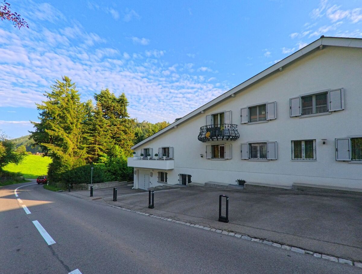 Ein langgestrecktes Wohnhaus mit hellen Fensterlaeden steht in Kehrsatz (3122, Kanton Bern) an einer leicht geschwungenen Strasse mit Blick auf eine gruene Wiese.