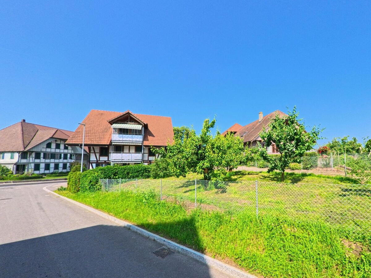 In Kernenried (3309, Kanton Bern) stehen mehrere traditionelle Häuser mit roten Ziegeldächern entlang einer kurvigen Straße, umgeben von grünen Wiesen und Bäumen.