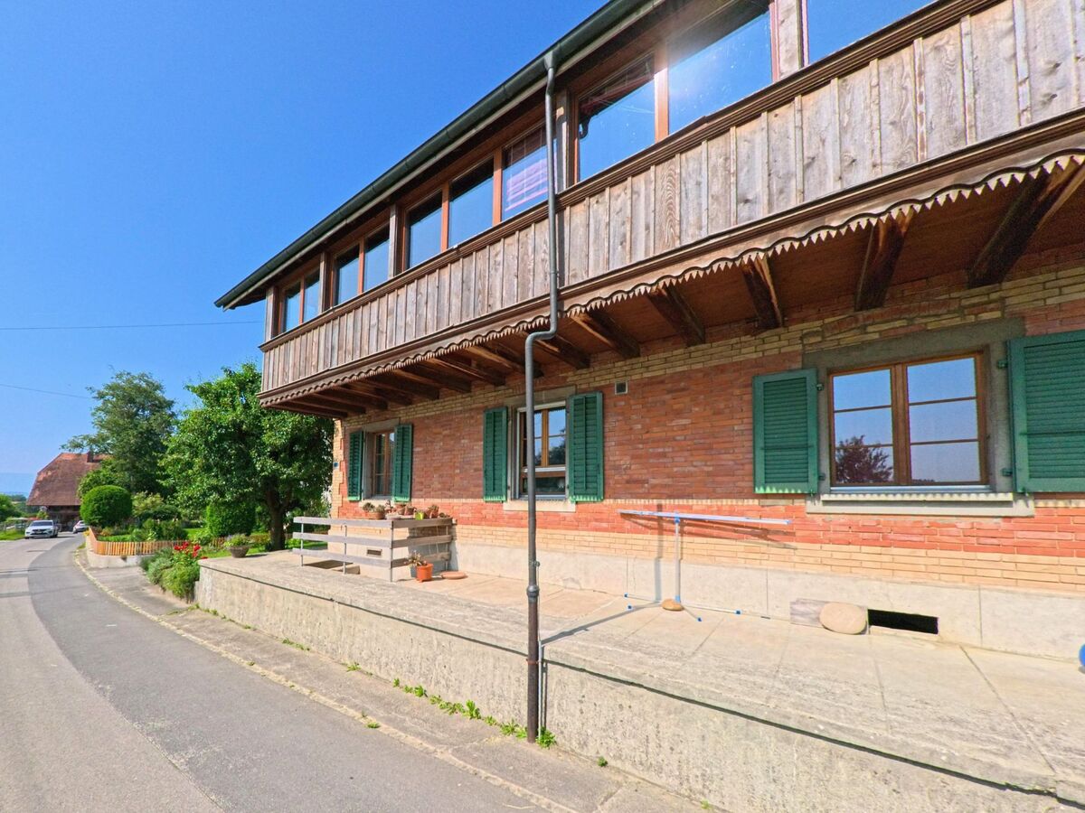 In Kernenried (3309, Kanton Bern) ist die Seite eines traditionellen Hauses mit einem Balkon und grünen Fensterläden zu sehen, das von einem gepflegten Garten und einer Straße umgeben ist.