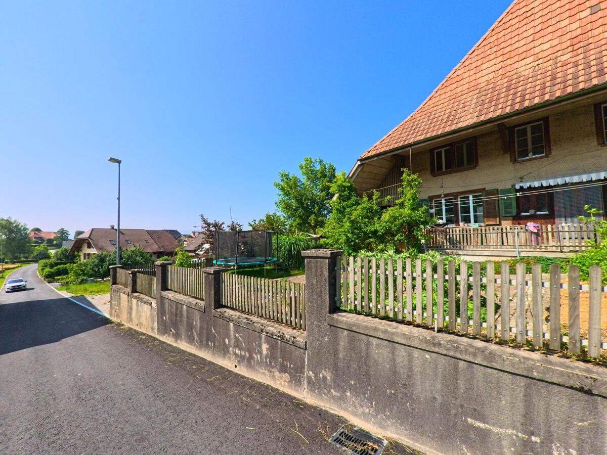 In Kernenried (3309, Kanton Bern) führt eine Straße an einem traditionellen Haus entlang, das von einem Holzzaun und einem Garten mit Trampolin umgeben ist.
