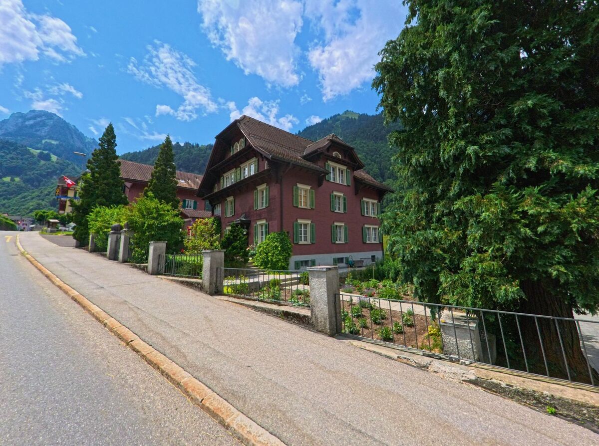 Ein grosses, rotes Wohnhaus mit grünen Fensterlaeden steht in Kerns (6067, Kanton Obwalden) am leicht ansteigenden Strassenrand, umgeben von Bäumen und einem gepflegten Vorgarten.