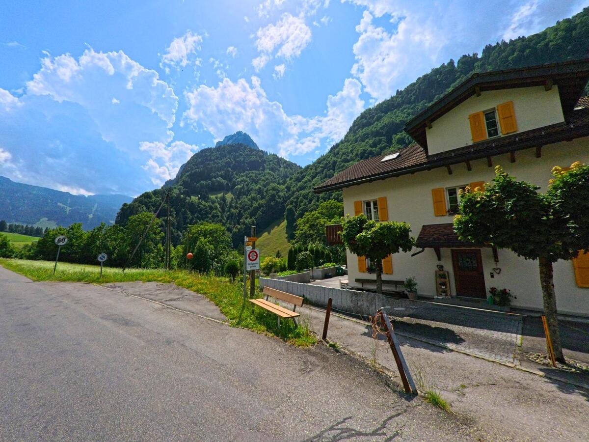 Am Ortsrand von Kerns (6067, Kanton Obwalden) steht ein helles Wohnhaus mit gelben Fensterlaeden neben einer ruhigen Strasse vor bewaldeten Huegeln.