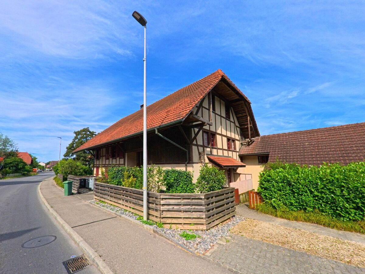 Entlang einer kurvigen Strasse in Kerzers (3207, Kanton Freiburg) steht ein gepflegtes Fachwerkhaus mit grosszügigem Vordach und angrenzendem Gartenzaun.