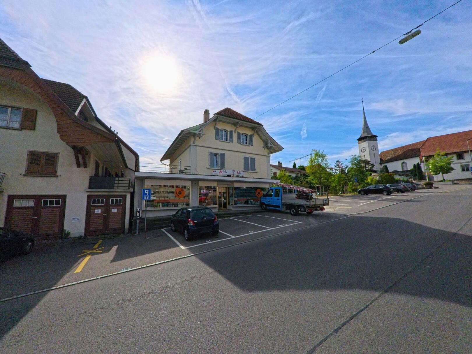 Im Zentrum von Kerzers (3207, Kanton Freiburg) zeigt sich ein kleiner Supermarkt mit Parkplätzen und der Kirche im Hintergrund bei sonnigem Wetter.