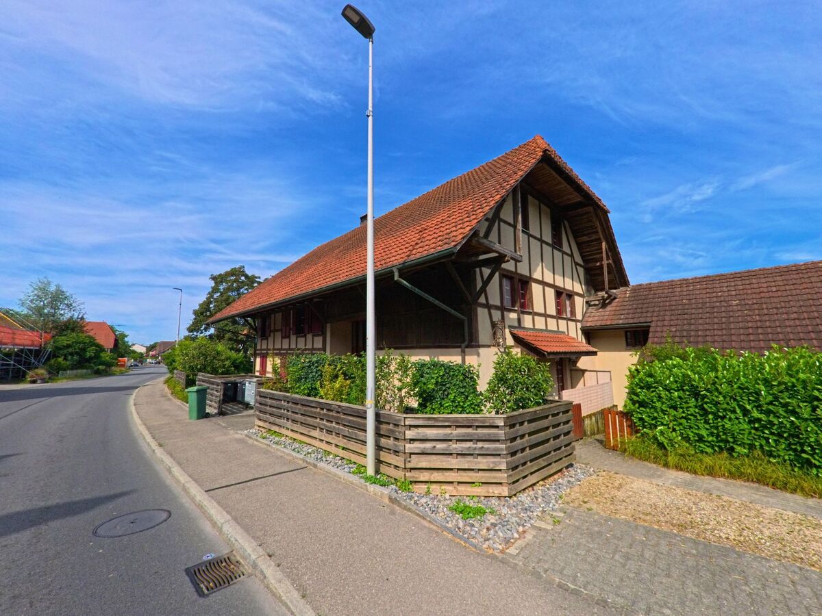 In Kerzers (3207, Kanton Freiburg) steht ein traditionelles Bauernhaus mit rotem Ziegeldach und Fachwerkfassade an einer leicht geschwungenen Strasse.