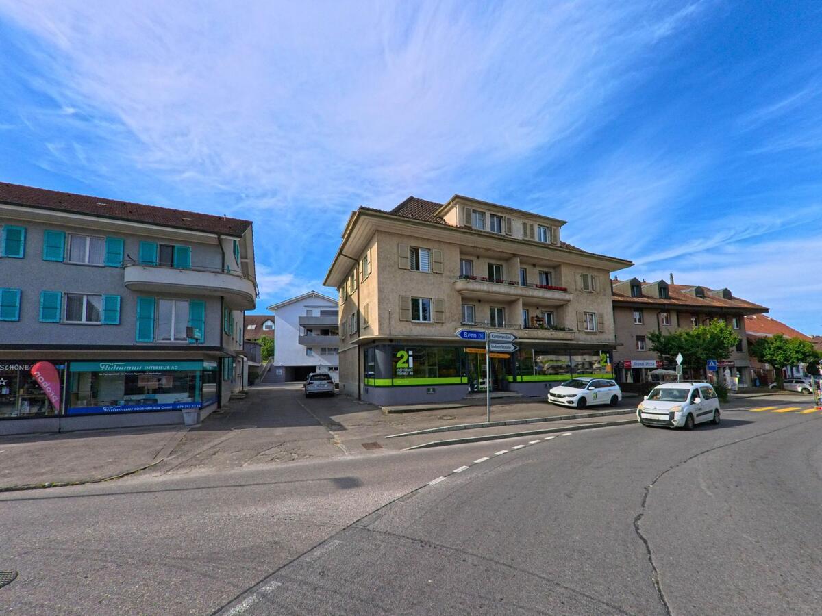 Im Zentrum von Kerzers (3207, Kanton Freiburg) prägen mehrere Geschäfts- und Wohnhäuser mit farbigen Fensterläden und Schaufenstern das Strassenbild an einer Kreuzung.