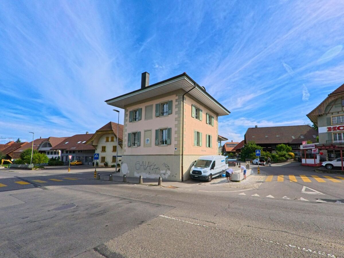 Direkt an einer Kreuzung im Zentrum von Kerzers (3207, Kanton Freiburg) steht ein helles Wohnhaus mit grünen Fensterläden und angrenzendem Zebrastreifen.