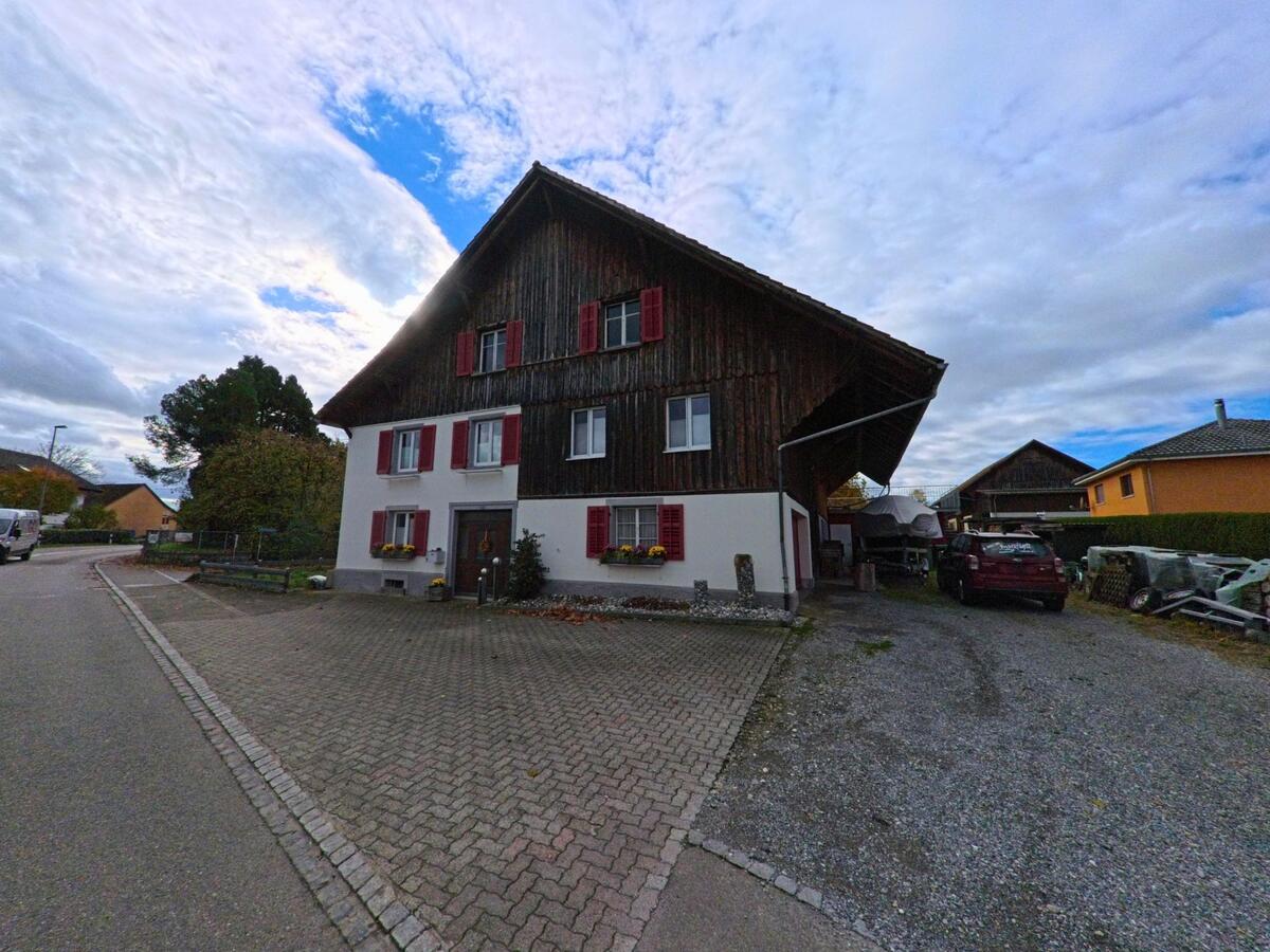 Ein grosses Bauernhaus mit dunkler Holzfassade und roten Fensterläden steht in Kesswil (8593, Kanton Thurgau) neben einem gepflasterten Vorplatz und einer Einfahrt mit Fahrzeugen.