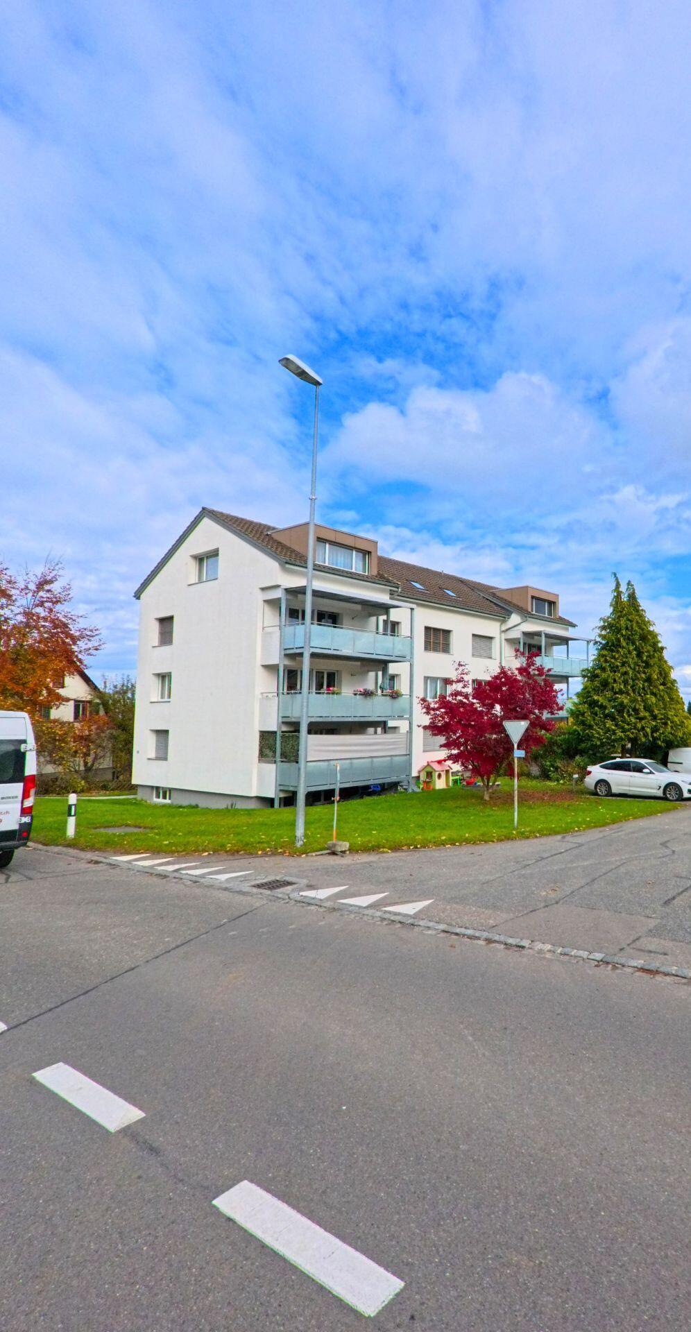 Ein modernes Mehrfamilienhaus mit Balkonen steht in Kesswil (8593, Kanton Thurgau) an einer ruhigen Kreuzung, umgeben von Rasenfläche und herbstlich gefärbten Bäumen.
