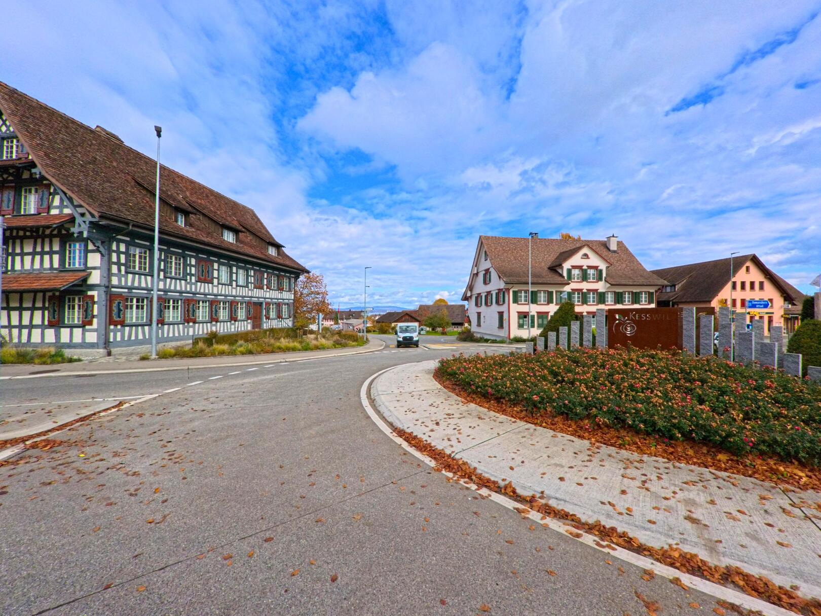 Am Ortseingang von Kesswil (8593, Kanton Thurgau) führt eine kurvige Strasse am bepflanzten Kreisverkehr mit Ortsschild vorbei, eingerahmt von traditionellen Wohnhäusern und einem Fachwerkbau.