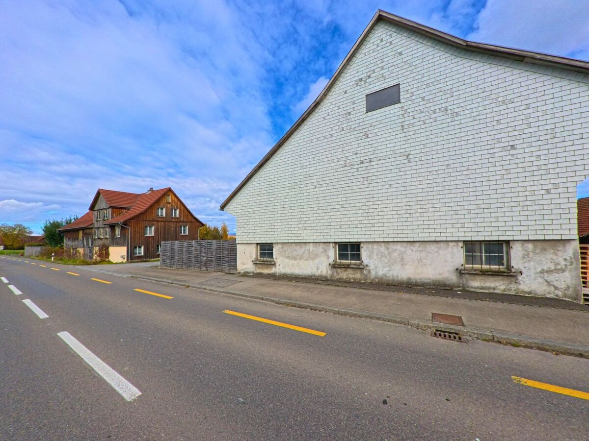 An einer geraden Strasse in Kesswil (8593, Kanton Thurgau) stehen ein grosses Gebäude mit heller Schindelfassade und ein Holzhaus mit rotem Ziegeldach am Rand des Quartiers.