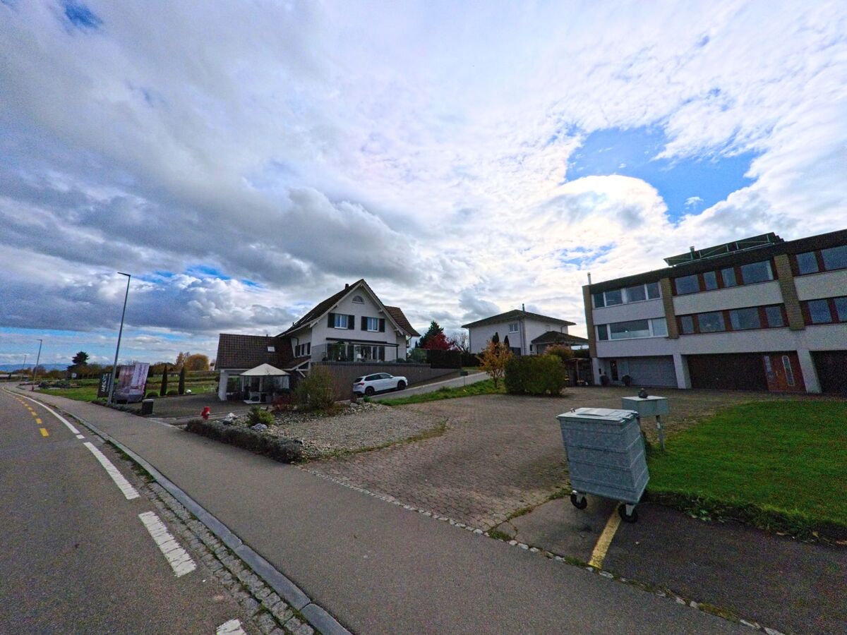 Am Rand einer Strasse in Kesswil (8593, Kanton Thurgau) stehen Wohnhäuser und ein grösseres Gebäude mit breiten Fensterbändern, davor ein Vorplatz mit Kies und einem rollbaren Container.