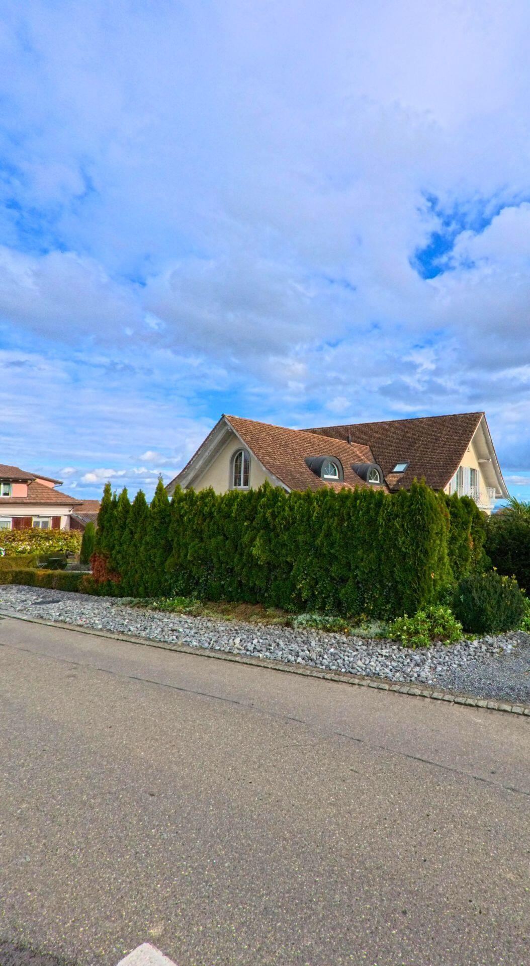 Hinter einer dichten Hecke zeigt sich in Kesswil (8593, Kanton Thurgau) ein Wohnhaus mit ziegelgedecktem Dach und Rundfenstern unter einem weitläufigen Himmel.