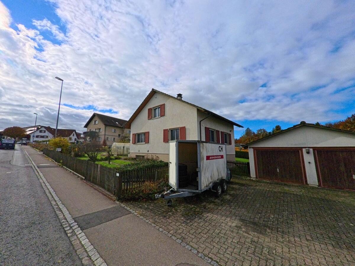 An einer Wohnstrasse in Kesswil (8593, Kanton Thurgau) steht ein helles Einfamilienhaus mit roten Fensterläden, neben einer Doppelgarage und einem abgestellten Anhänger auf dem Vorplatz.