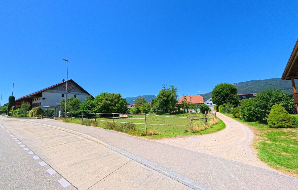 In Kestenholz (4703, Kanton Solothurn) verläuft ein unbefestigter Weg zwischen Wiesen und Bauernhäusern, im Hintergrund erheben sich bewaldete Hügel.