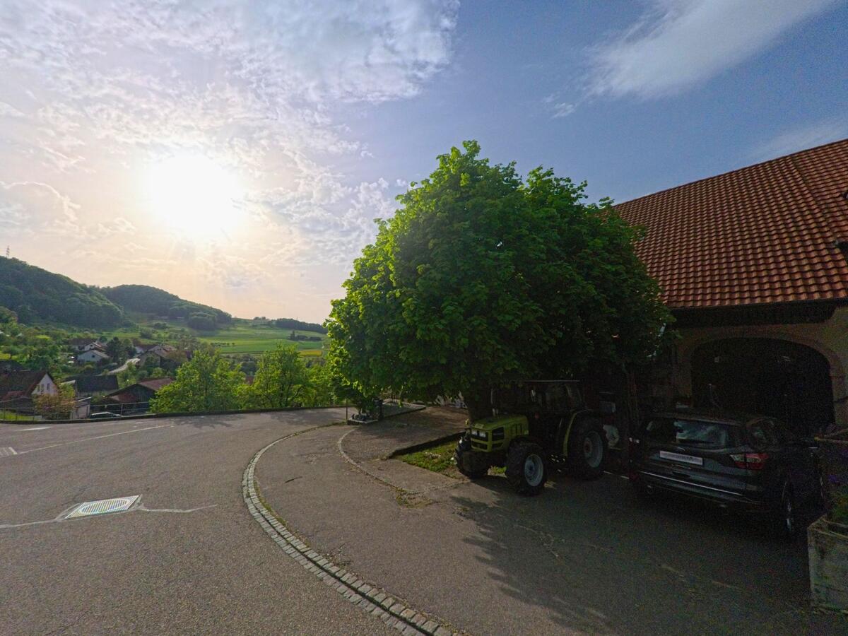 Am Ortsrand von Kienberg (4468, Kanton Solothurn) steht ein Bauernhof mit Traktor unter einem grossen Baum, dahinter öffnet sich der Blick ins Tal.