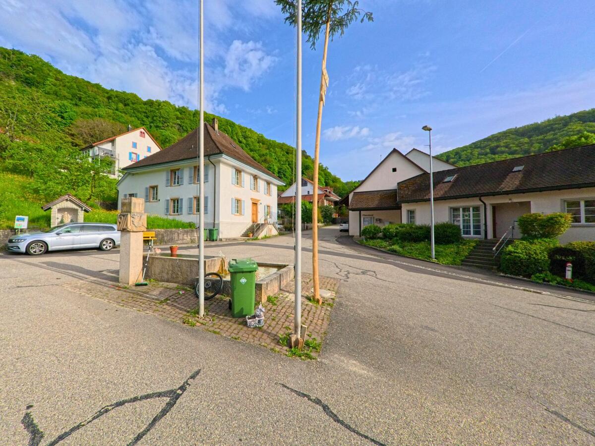 Im Zentrum von Kienberg (4468, Kanton Solothurn) stehen historische und moderne Gebäude nebeneinander, mit Brunnenanlage und Fahnenmast auf dem Platz.