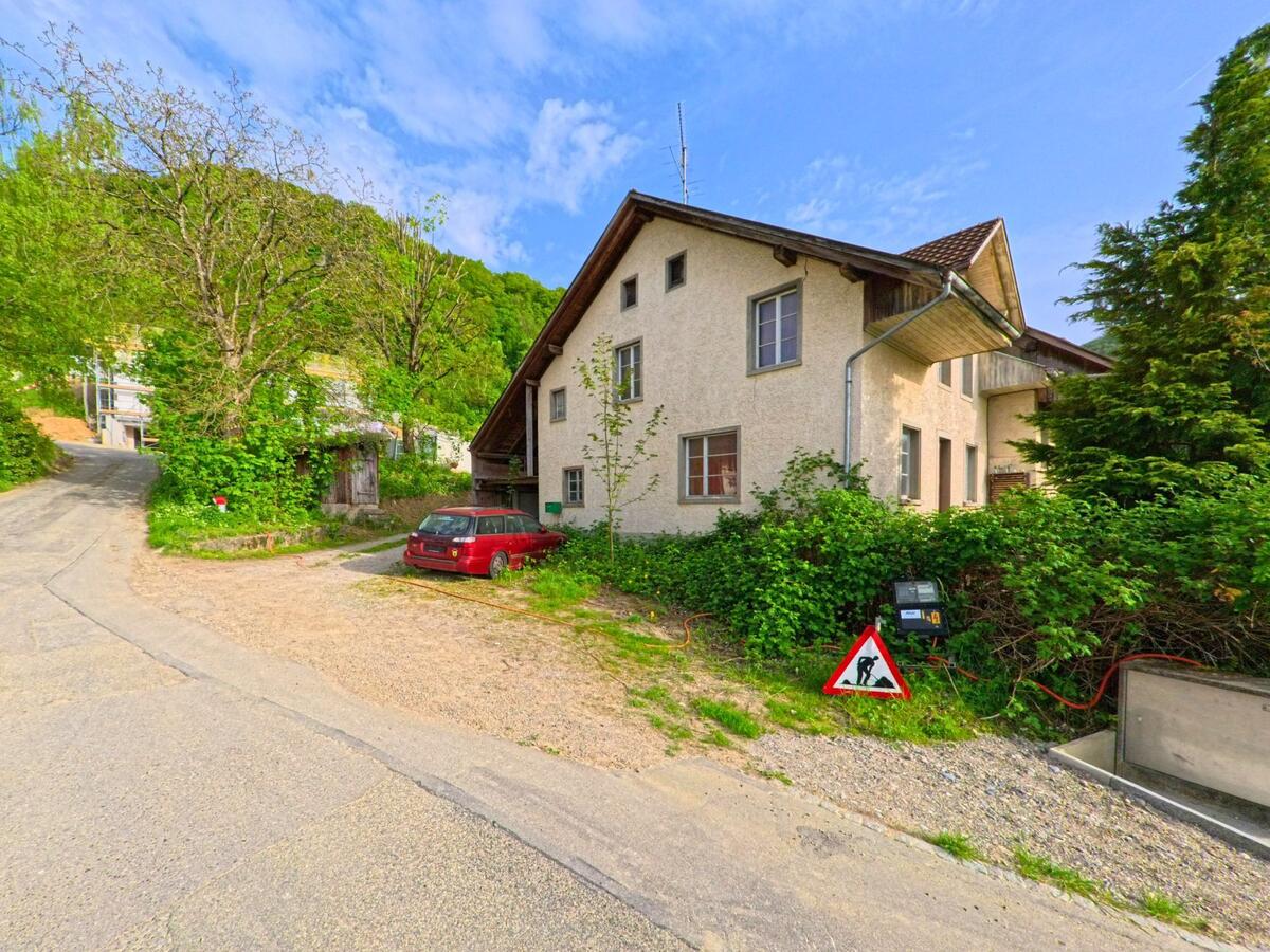 Am Rand einer ruhigen Strasse in Kienberg (4468, Kanton Solothurn) steht ein älteres Wohnhaus mit Kiesparkplatz und Bauhinweis zwischen Bäumen.