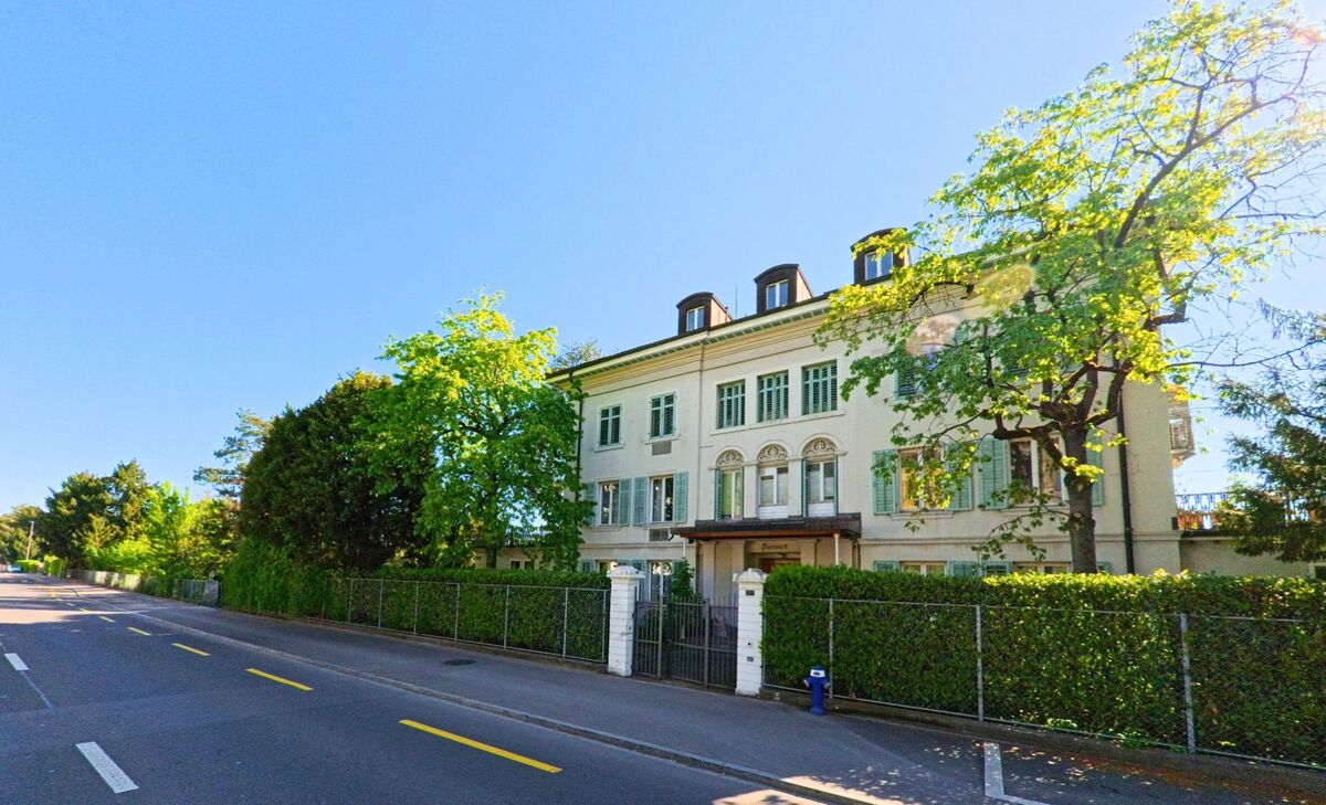 In Kilchberg (8802, Kanton Zürich) steht ein herrschaftliches Gebäude mit grünen Fensterläden und verzierten Fassadenelementen hinter einer Hecke an einer ruhigen Strasse.
