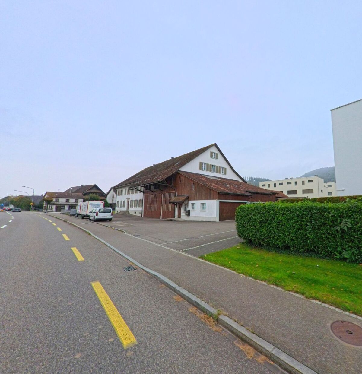 Ein traditionelles Bauernhaus mit Holzfassade und roten Ziegeldach in Killwangen (8956, Kanton Aargau) steht an einer Hauptstrasse mit gelber Markierung und angrenzenden Neubauten.