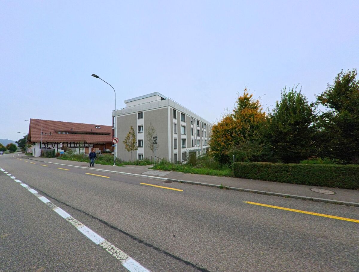 An der Hauptstrasse in Killwangen (8956, Kanton Aargau) stehen ein modernes Mehrfamilienhaus und ein traditionelles Fachwerkhaus nebeneinander, getrennt durch eine Baumreihe.