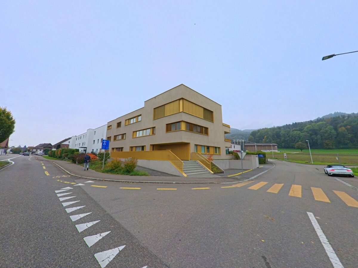 Ein modernes Gebäude mit gelben Fensterläden und Treppenaufgang steht in Killwangen (8956, Kanton Aargau) an einer Strassenkreuzung mit Fussgängerstreifen und Radweg.