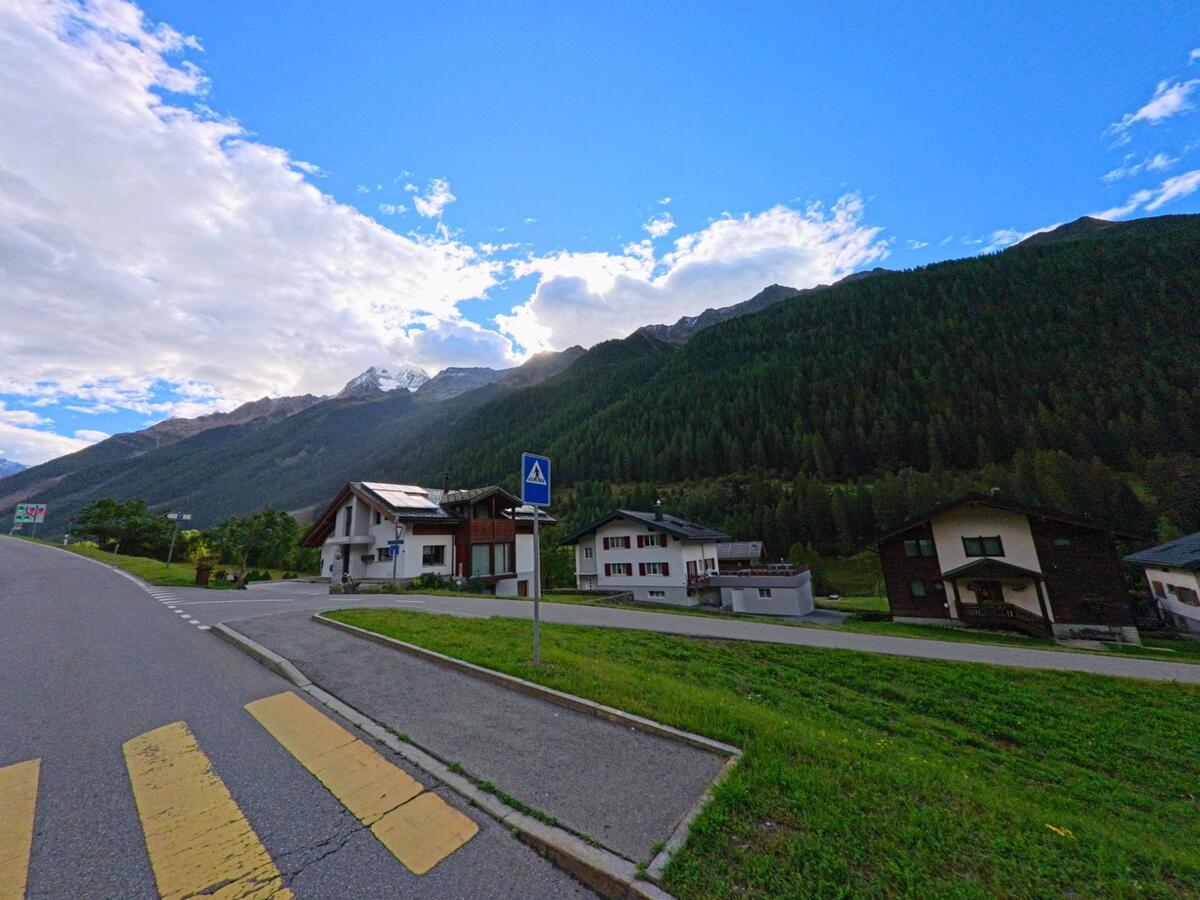 Ein Fussgaengerstreifen fuehrt zu Wohnhaeusern am Ortsrand von Kippel (3917, Kanton Wallis), im Hintergrund ragen bewaldete Berge mit schneebedeckten Gipfeln auf.