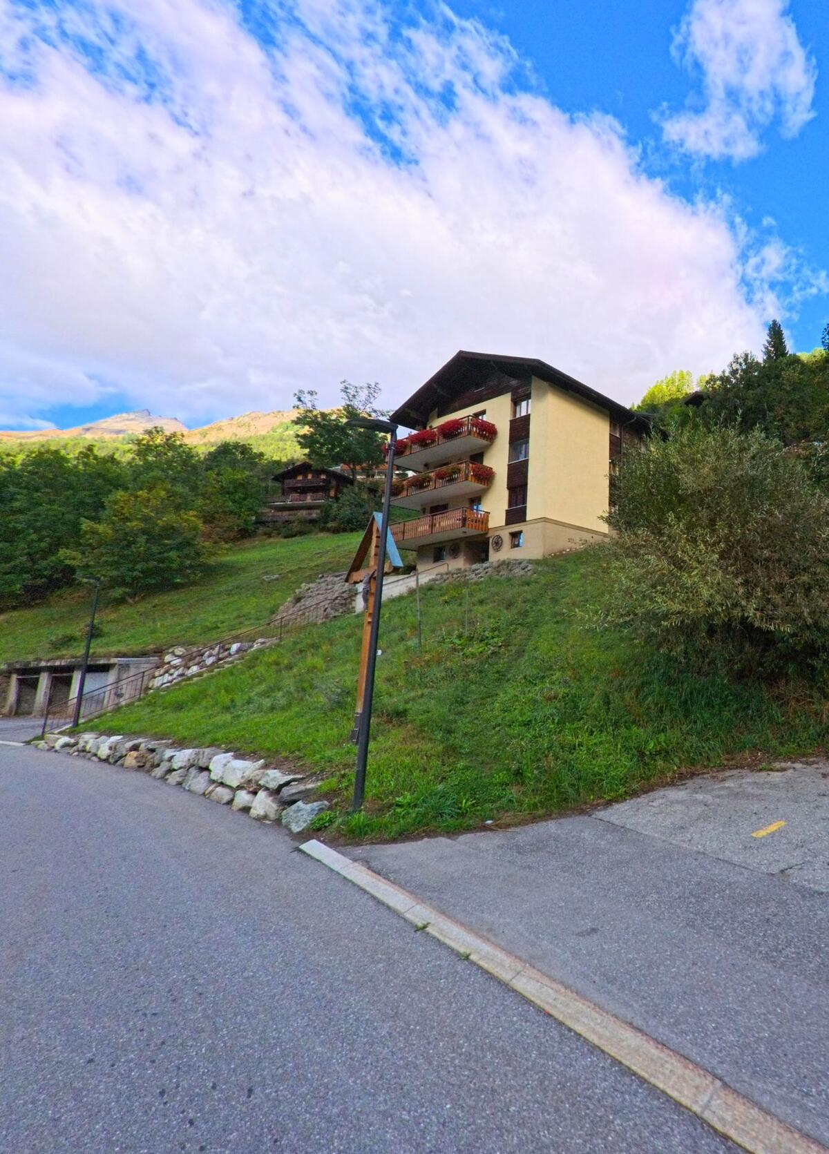 Ein gelbes Mehrfamilienhaus mit Balkonen und Blumenschmuck erhebt sich am Hang in Kippel (3917, Kanton Wallis) vor bewaldeten Bergen.
