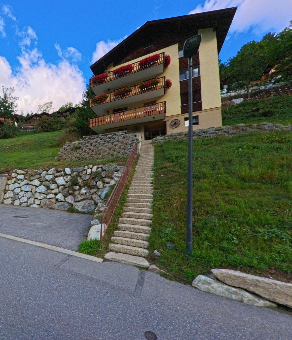 Ein Mehrfamilienhaus in Kippel (3917, Kanton Wallis) mit Balkonen voller Blumenschmuck ist ueber eine lange Steintreppe erreichbar.