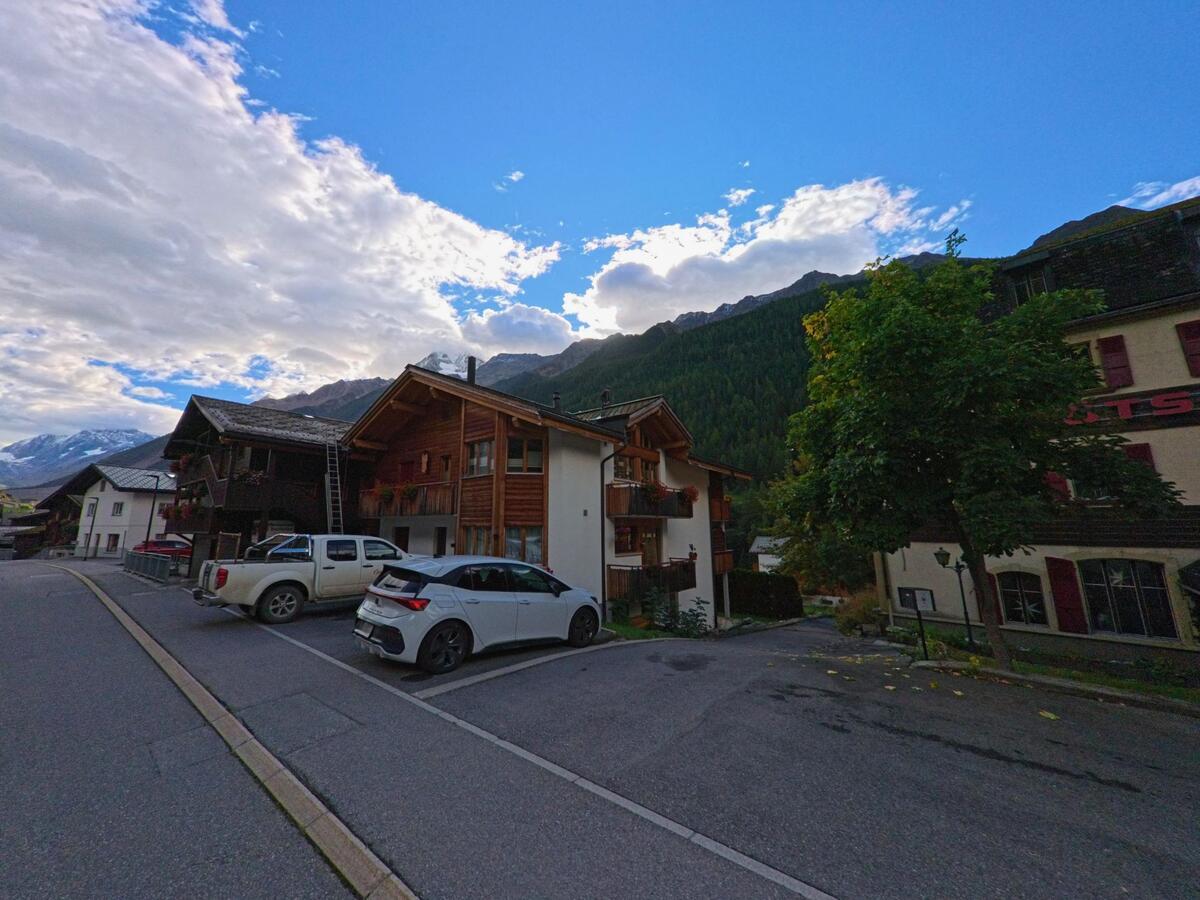 In Kippel (3917, Kanton Wallis) reihen sich Wohnhaeuser und Chalets entlang einer Strasse, im Hintergrund sind bewaldete Berge und schneebedeckte Gipfel sichtbar.