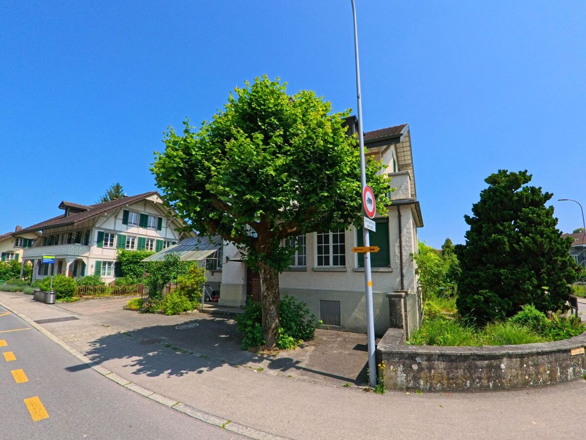 In Kirchberg (3422, Kanton Bern) säumen gepflegte Häuser mit grünen Fensterläden und Vorgärten eine ruhige Strasse mit Baumallee.