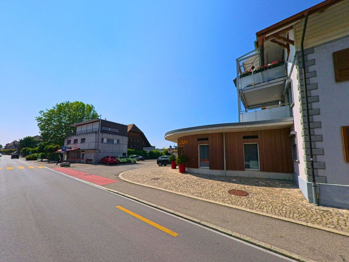 In Kirchberg (3422, Kanton Bern) liegt an einer Kreuzung ein modernes Gebäude mit Holzverkleidung neben einem älteren Wohn- und Geschäftshaus mit Vorplatz.